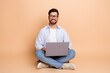 © deagreez - Positive young man sitting on beige background with a laptop showcasing attractiveness and leisure in stylish casual attire