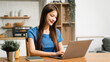 © NanSan - An Asian woman sits comfortably at her home desk, focused on her laptop. She works online in a relaxed setting, managing tasks efficiently with dedication, concentration, and a positive attitude