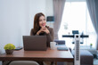 © Tj - Young Asian businesswoman smiling and looking away while working from home at a wooden table with a laptop, smartphone, tablet, coffee cup, and plant