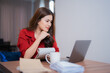 © Tj - Young asian businesswoman working from home, sitting at wooden table and using laptop, taking notes on notepad, holding pen and thinking, with cup of coffee and documents nearby