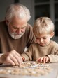 © Irina - Elderly man and a young boy sitting at a table and playing with a jigsaw puzzle. the man is on the left side of the image, with a white beard and is wearing a brown sweater.