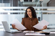 © Liubomir - A concerned businesswoman reviews documents at her desk, surrounded by office supplies and technology. She looks perplexed, possibly due to the content.