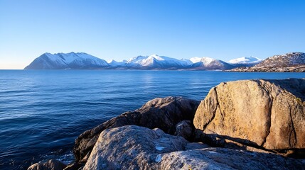 Poster - Serene coastal landscape featuring snow-capped mountains reflected in a calm, blue ocean, with large rocks in the foreground under a clear sky. : Generative AI