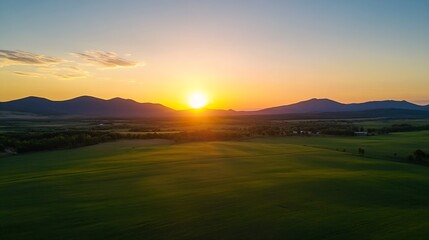 Poster - Aerial view of a vibrant green field at sunset, with silhouetted mountains in the background and a warm, golden sky. : Generative AI