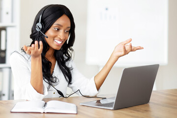 Aufkleber - Smiling entrepreneur or office worker in headset speaking with business partner, having online videocall in office. Millennial black secretary communicating on webcam, using laptop computer