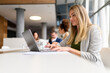 © Moon Safari - Focused blond businesswoman analyzing data over laptop while colleagues collaborating in the background in board room