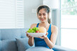 © M+Isolation+Photo - Asian woman in sportswear sits on a sofa, enjoying a fresh salad near her laptop. She focuses on healthy eating and wellness while managing her nutrition and lifestyle comfortably at home