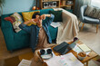 © DragonImages - Young man taking a break from work while sitting on a cozy couch in a living room filled with papers, books, and laptop. Relaxed home office setup with various gadgets and accessories