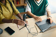 © DragonImages - Two colleagues setting up a microphone for podcasting with an open laptop on desk. Various digital gadgets placed on table creating functional workspace