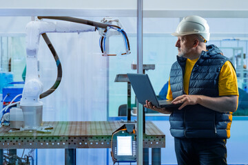 Poster - Engineer in hard hat programs robotic arm with laptop in high-tech factory environment, focusing on automation, precision, and modern industrial technology. Manufacturing.