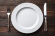 © Cheewynn - A white plate with a fork and knife on a wooden table, ready for a meal.