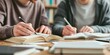 © Planetz - Two people are studying together at a table, writing notes in open books in a library or study space.