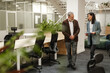 © Drobot Dean - A middle-aged man and woman are walking and discussing documents in a modern open-plan office. They are business partners engaged in a professional conversation near workstations.