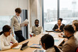 © Drobot Dean - A group of diverse coworkers meet in a modern conference room. A young man stands, holding documents, while the others sit around the table engaged in discussion.