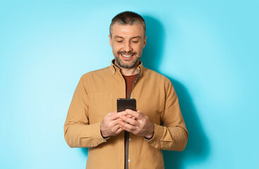 Poster - Cheerful Man Using Mobile Application On Cellphone Or Texting Via Smartphone Communicating Online Standing In Studio Over Blue Background. Communication And Technology