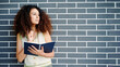 © khmelev - thoughtful woman holding notebook while standing against brick wall, pondering ideas and inspiration. casual fashion and curly hair emphasize creativity and contemplation