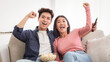 © Prostock-studio - Excited Chinese Couple Watching TV Shaking Fists In Joy Celebrating Victory Of Favorite Sport Team Sitting On Couch In Living Room At Home. Weekend Leisure, Television Show And Entertainment Concept