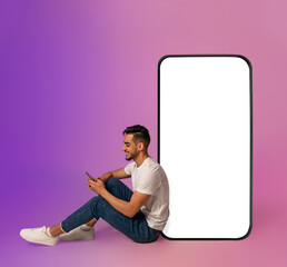 Poster - Cheery millennial Arab guy sitting near big cellphone with blank white screen, using mobile device, using new app in neon light, mockup for website or application design