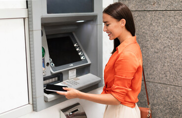 Young lady customer using smartphone to gain access to ATM, withdrawing money from account standing near bank machine on a street outside, side view shot. Financial safety and transactions