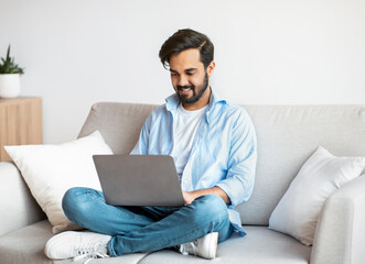 Poster - Distant Work. Happy Arab Freelancer Man Using Laptop Computer At Home, Typing On Keyboard, Young Eastern Guy Sitting On Sofa In Living Room, Working Remotely On Online Project, Copy Space