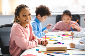 Poster - Academic Concept. Smiling junior african american school girl sitting at desk in classroom, writing in notebook, posing and looking at camera. Group of classmates studying in the background