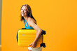 © SHOTPRIME STUDIO - Cheerful woman holding a vibrant yellow suitcase standing against a bold orange wall with a joyful expression
