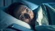© Rona_65 - A woman lies in bed at night, eyes wide open in fear, illuminated by blue light with a phone beside her on the pillow.
