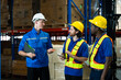 © Ekkasit A Siam - Logistics team of three male workers wearing safety helmets and vests engaged in discussion inside warehouse. One holding clipboard, others holding tablet and paper, with boxes stacked behind.
