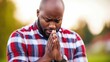 © imagineRbc - A man is praying in a park. He is wearing a red and white plaid shirt. The man is looking down and his hands are clasped together
