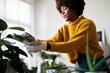 © Studio Marmellata - A woman wearing gloves gently cleans the leaves of her houseplant. She is wearing a yellow sweater and jeans in a bright, modern room.
