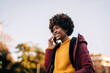 © Studio Marmellata - A young woman with an afro is outside, talking on her phone. She is smiling and wearing a yellow sweater and a maroon jacket.