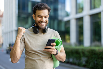 Close-up photo of a young smiling Indian man wearing headphones and a mat, looking at the phone screen and showing a victory gesture with his hand