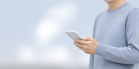 Poster - Man holding smartphone in casual blue sweater, standing against soft blurred background, showcasing modern technology and communication in a serene environment