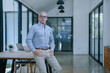 © Tj - Confident mature businessman wearing eyeglasses and a blue shirt, standing in a modern office, with hands in pockets and leaning on a desk featuring a laptop, exuding professionalism