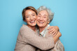 © ABBY - Smiling grandmother and granddaughter kissing cheeks in front of a bright blue background. A joyful family scene celebrating Mother’s Day and generational love.