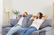 © Studio Romantic - Smiling young family couple relaxing while sitting with hands behind their heads on couch in living room at home. Wife and husband resting together with satisfied expression, enjoying peaceful break.