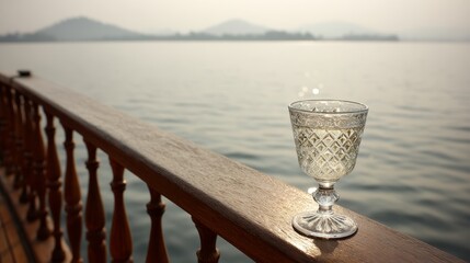 Fototapete - A crystal wine glass sits elegantly on a wooden yacht railing, illuminated by warm sunset light reflecting on distant misty hills