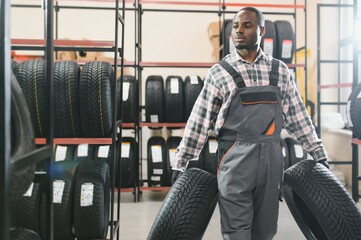 Poster - Car tire service concept. Portrait African american man mechanic holding new tyre on background store