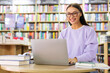 © Home-stock - Doing research. Happy European woman in glasses using laptop computer in library, smiling at screen while studying