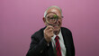 © Krakenimages.com - Senior man in business attire holds magnifying glass against isolated pink background suggestive of investigation, observation, and analytical concepts.