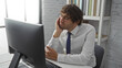 © Krakenimages.com - Young man in office chair looking tired at computer, wearing white shirt and tie, surrounded by bookshelves, daylight through window highlighting focused expression.