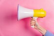 © AR - A hand holds a vibrant yellow and white megaphone against a pink background perfect for announcement promotion and marketing concepts.
