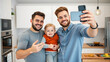 © Alexandra - Young male gay couple taking a selfie on a smartphone with their son in the kitchen of their new apartment