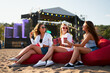 © artiemedvedev - Girls sitting on red bean bag chairs, clinking drinks at beach music festival. Sunglasses, casual summer outfits, sea stage, relaxed vibe. Friends enjoy concert, sunny day, refreshing beverages.