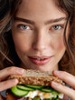 © Nick Freund - Nahaufnahme einer jungen Frau mit natürlichem Makeup, die in die Kamera schaut und ein Sandwich hält
