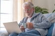 © luciano - Senior relaxed smiling man sitting on sofa browsing internet on laptop holding a coffee cup - indoor, at home concept - caucasian elderly retired man using technology