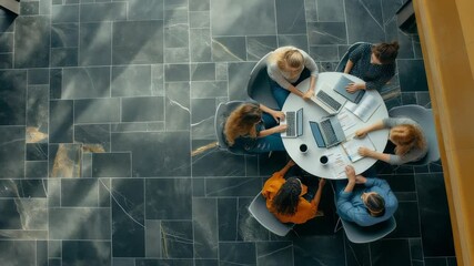 Poster - A top-down view shows a group of six diverse individuals collaborating around a white round table with laptops and papers in a workspace featuring grey stone tiled flooring. Focused teamwork.