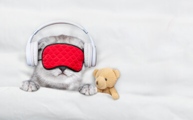 Leinwandbilder - Cute kitten wearing  headphones and mask for sleep listening music with toy bear on a bed at home. Top down view. Empty space for text