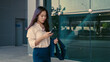 © Lifehdfilm - Asian office employee walking past glass building while using smartphone and holding blazer. Casual moment outside workplace during workday.