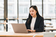 © kenchiro168 - Young Asian businesswoman in black suit working on laptop at modern office, focused and professional, natural light from large windows, coffee cup on desk, productivity concept